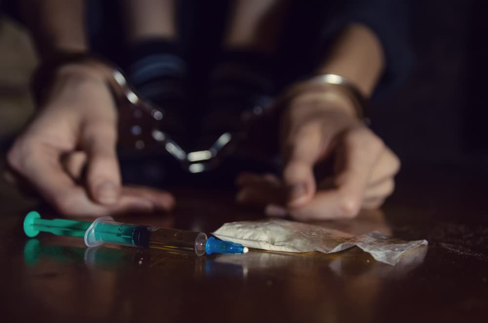 Syringe and drugs with a defocused man sitting on the floor and his hands locked in handcuffs.
